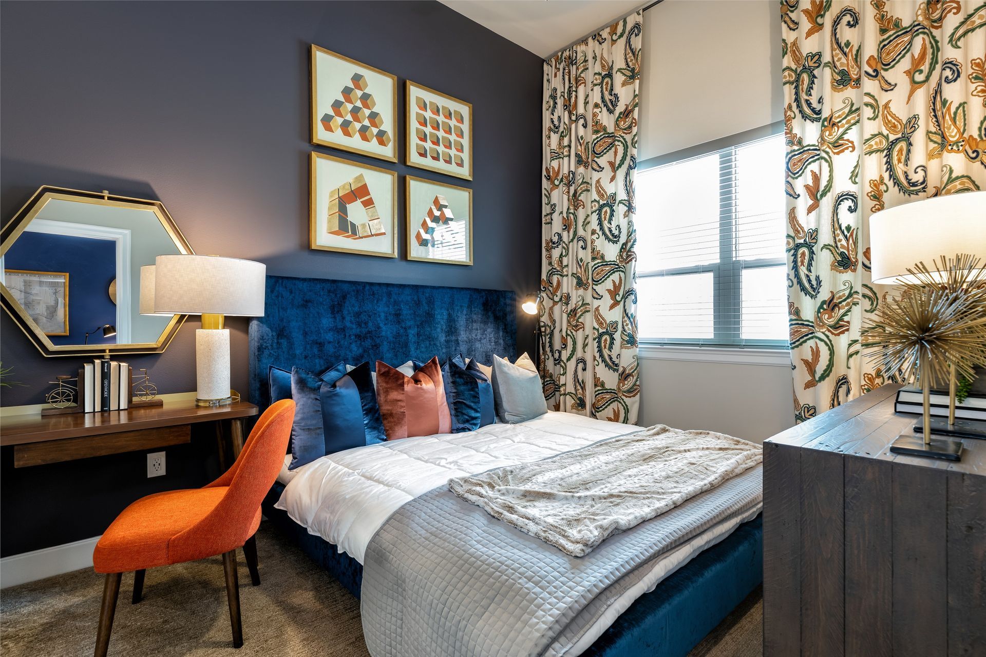 A bedroom with a blue velvet bed, orange chair, and decorative wall art. Features rich colors and patterns, with a desk and window.