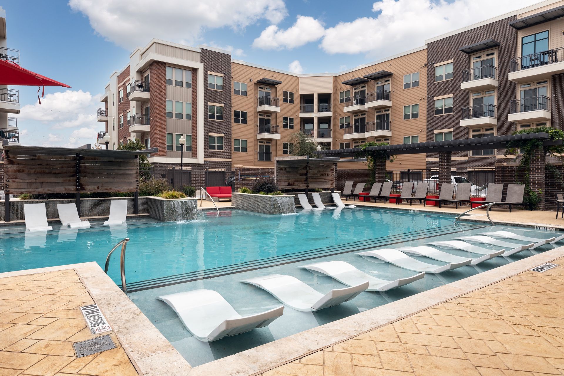 Apartment complex with a large swimming pool featuring in-pool lounge chairs. Buildings have brick and tan facades; the sky is blue with clouds.