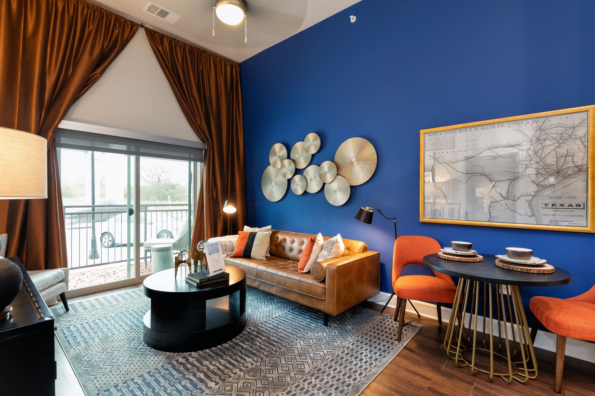 Living room with a blue wall, leather sofa, orange chairs, and a round table. Includes art and curtains.