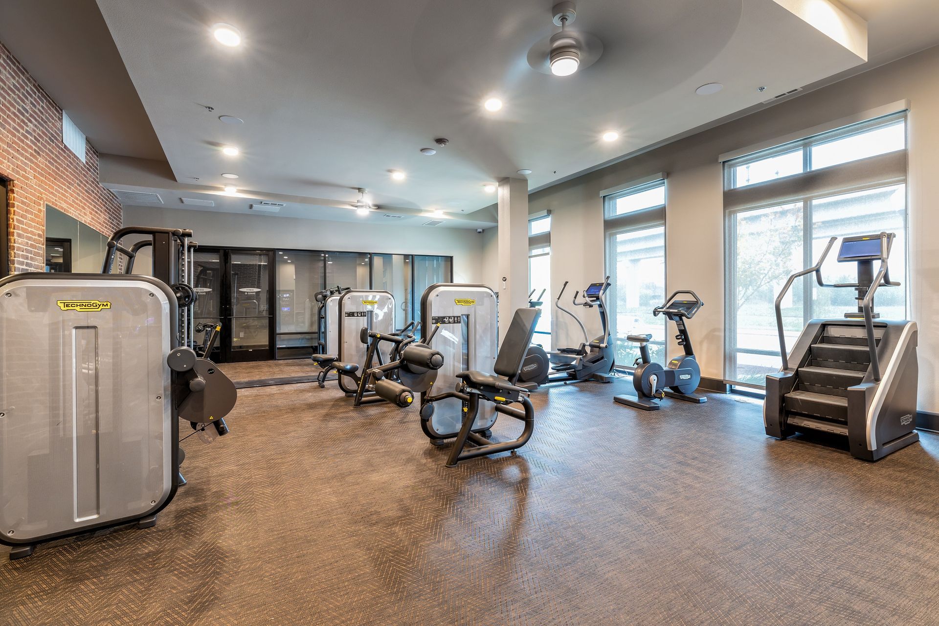A well-lit gym with various exercise machines on a speckled floor. Large windows let in natural light.