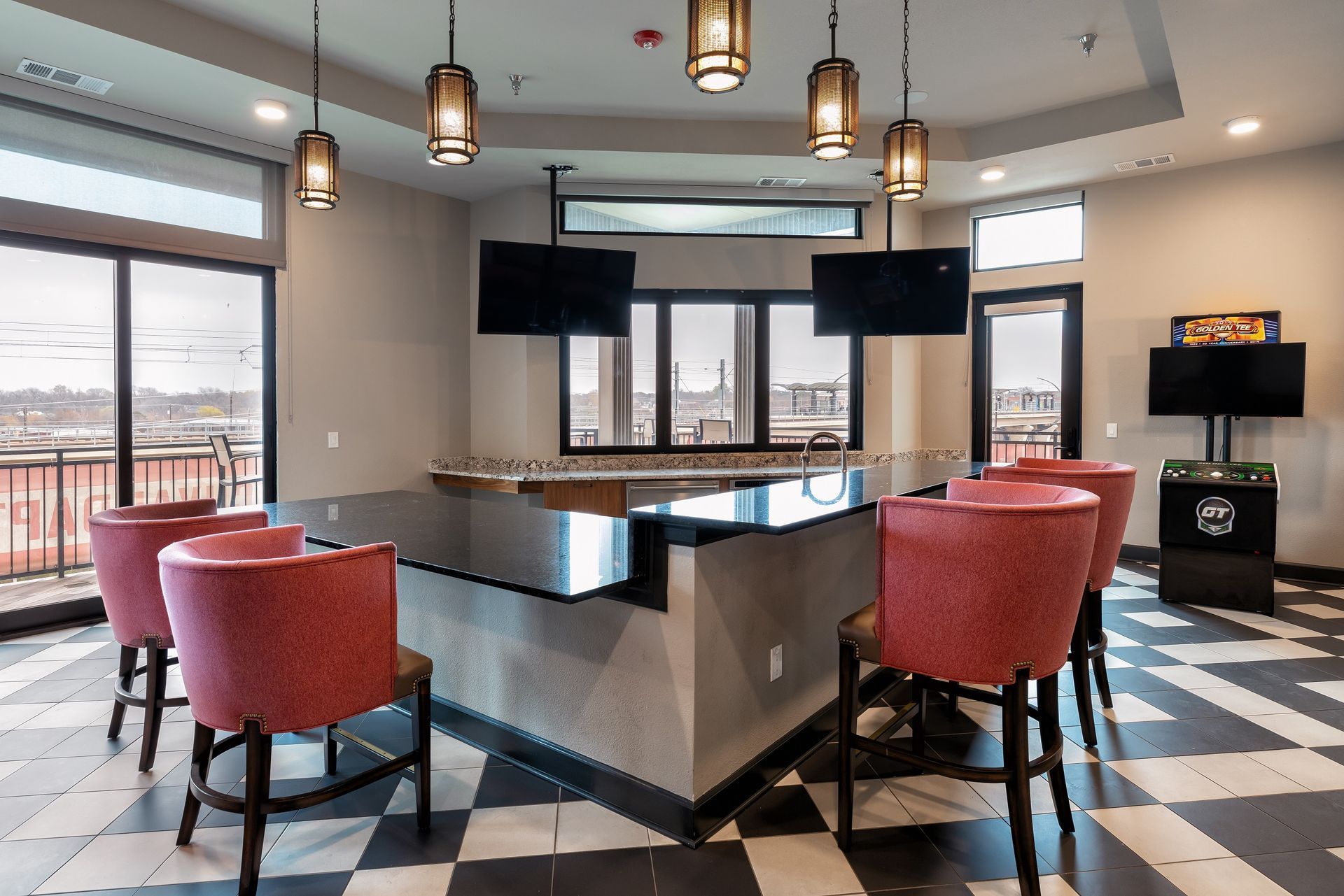 A bar area with red chairs, black countertops, and two mounted TVs. Features include hanging lights, windows, and a black and white tiled floor.