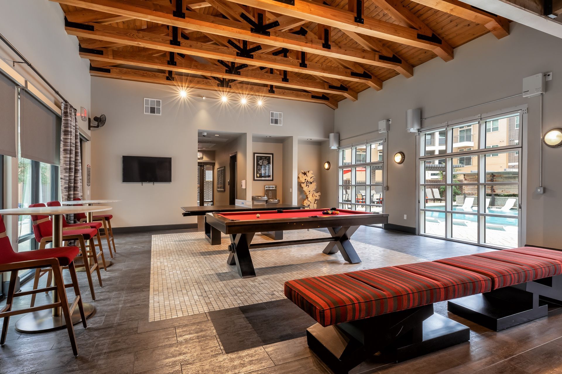 A well-lit recreation room with a pool table, red bar stools, a bench, and windows overlooking a pool.