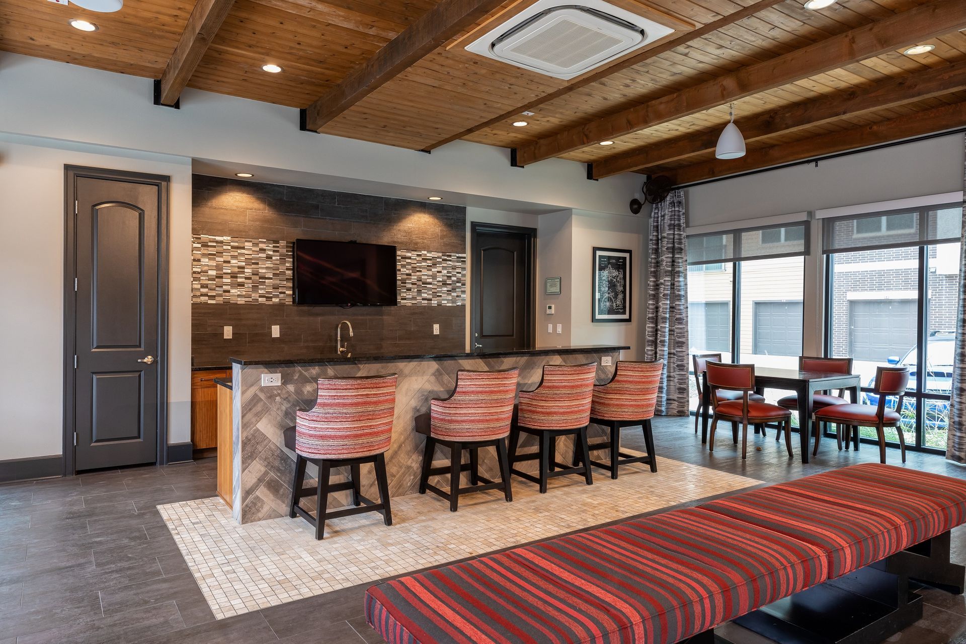 A lounge area with a bar, dining table, and bench seating. Features wood-beam ceiling, mosaic tile backsplash, and patterned bar stools.