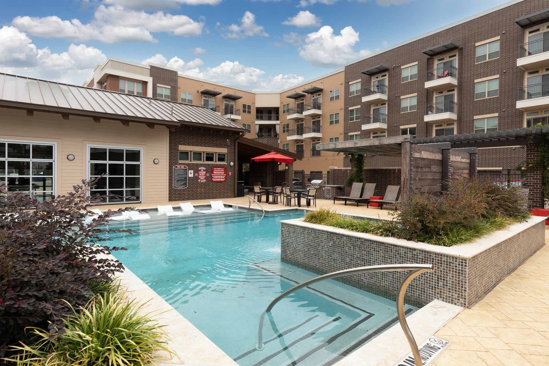Pool and patio area at an apartment complex, with buildings in the background. Pool is surrounded by lounge chairs, a grill area and a building with large windows.