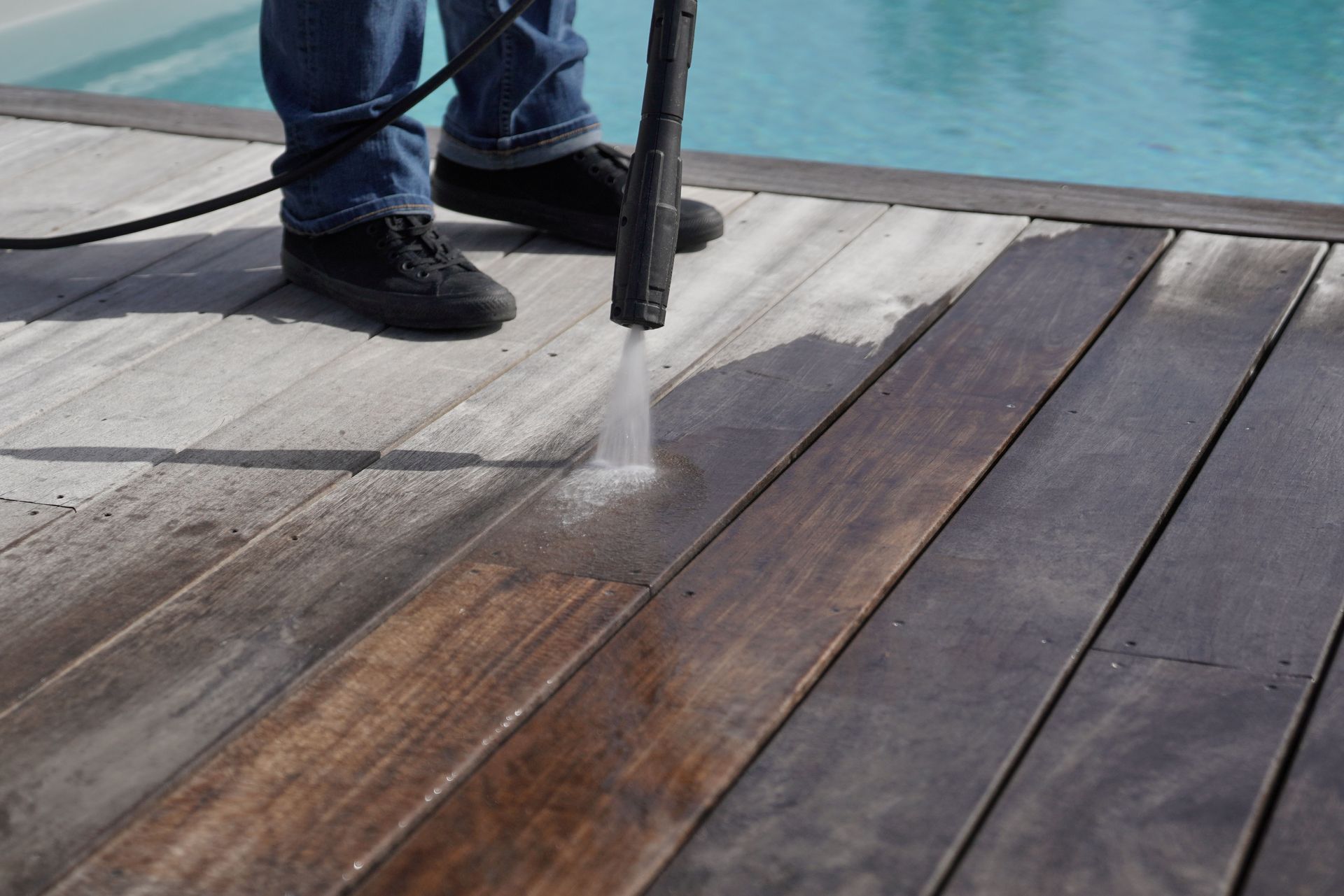 Person pressure-washes a wooden deck beside a pool.