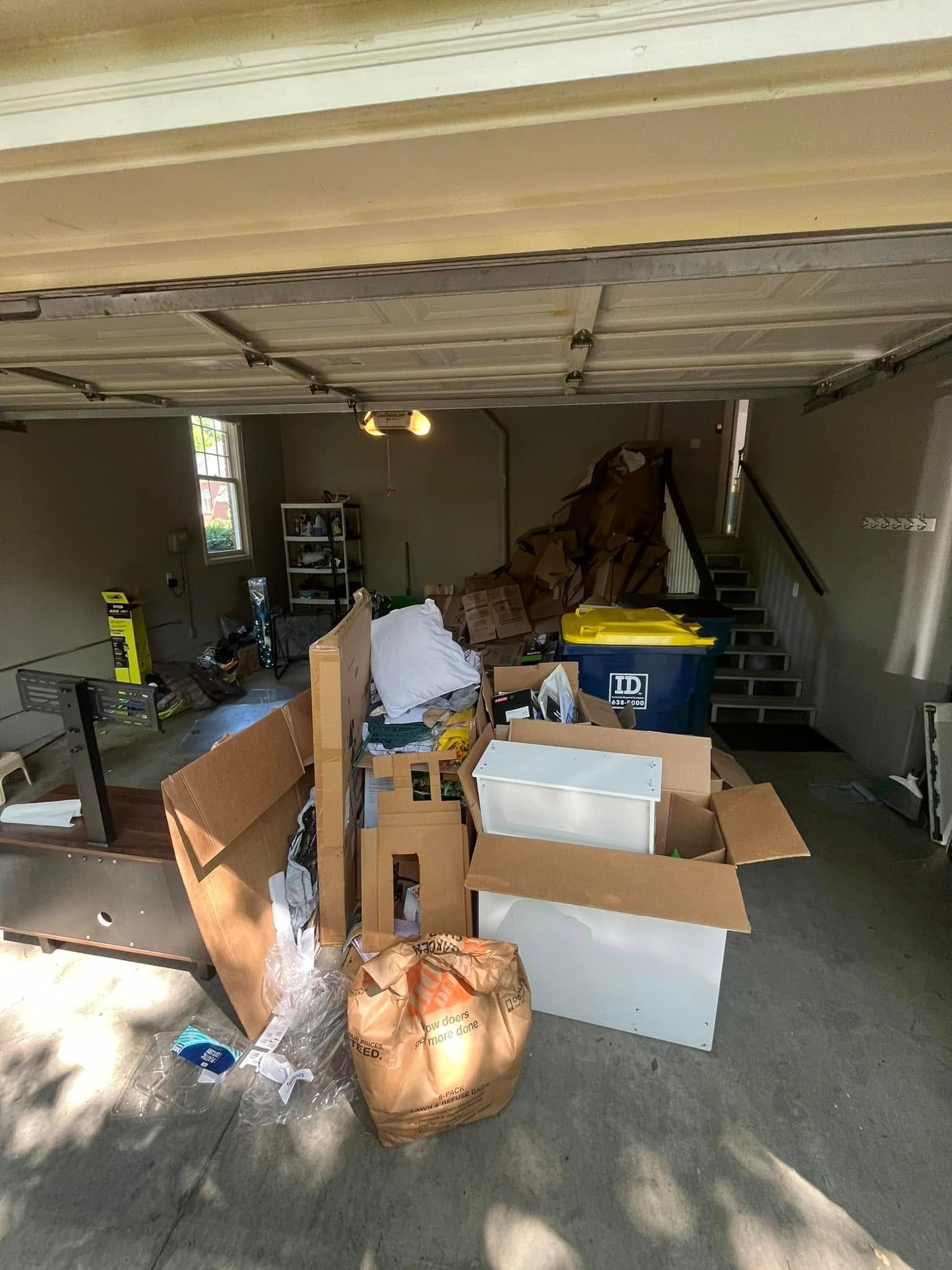 Cluttered garage with boxes, trash bags, and a yellow container; stairs in the background.