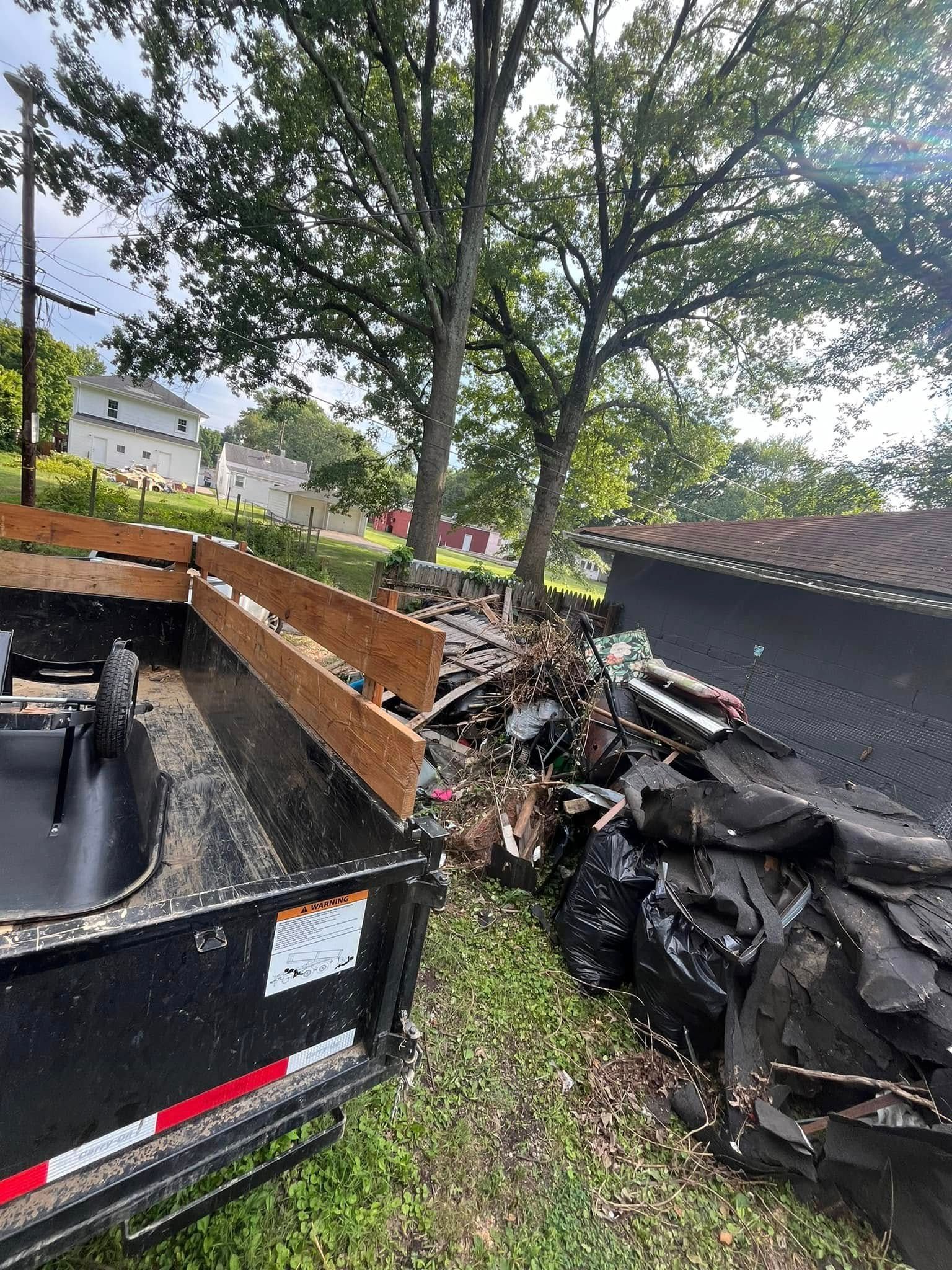Trailer filled with debris next to a pile of refuse and trees, outdoors.