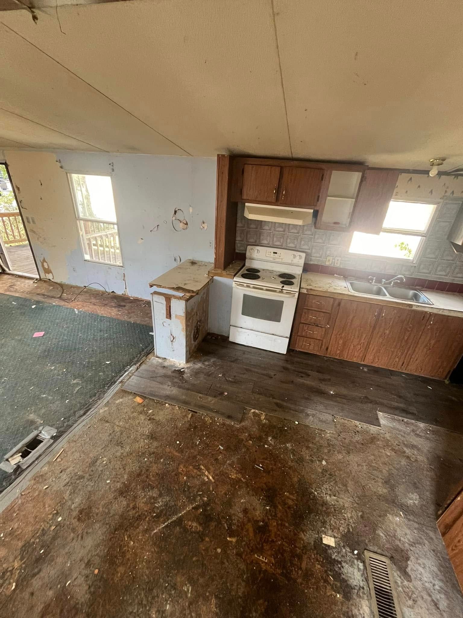Interior kitchen view in disrepair. White stove, damaged cabinets and walls, peeling flooring, and two windows.