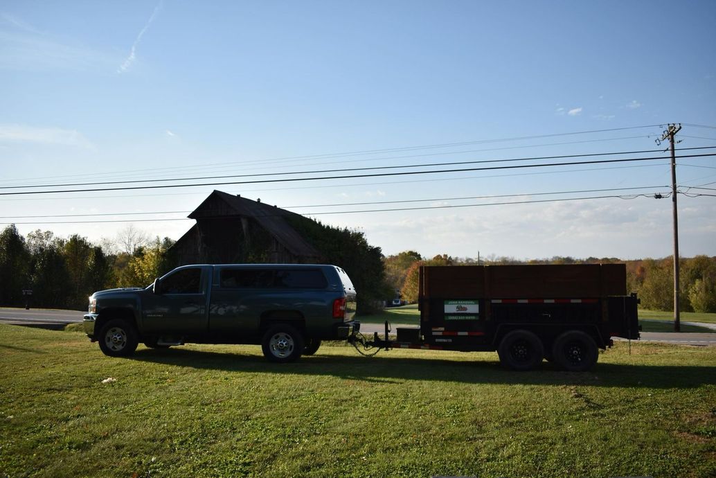 Blue truck towing a trailer on a grassy area, barn in the background. Sunny day.