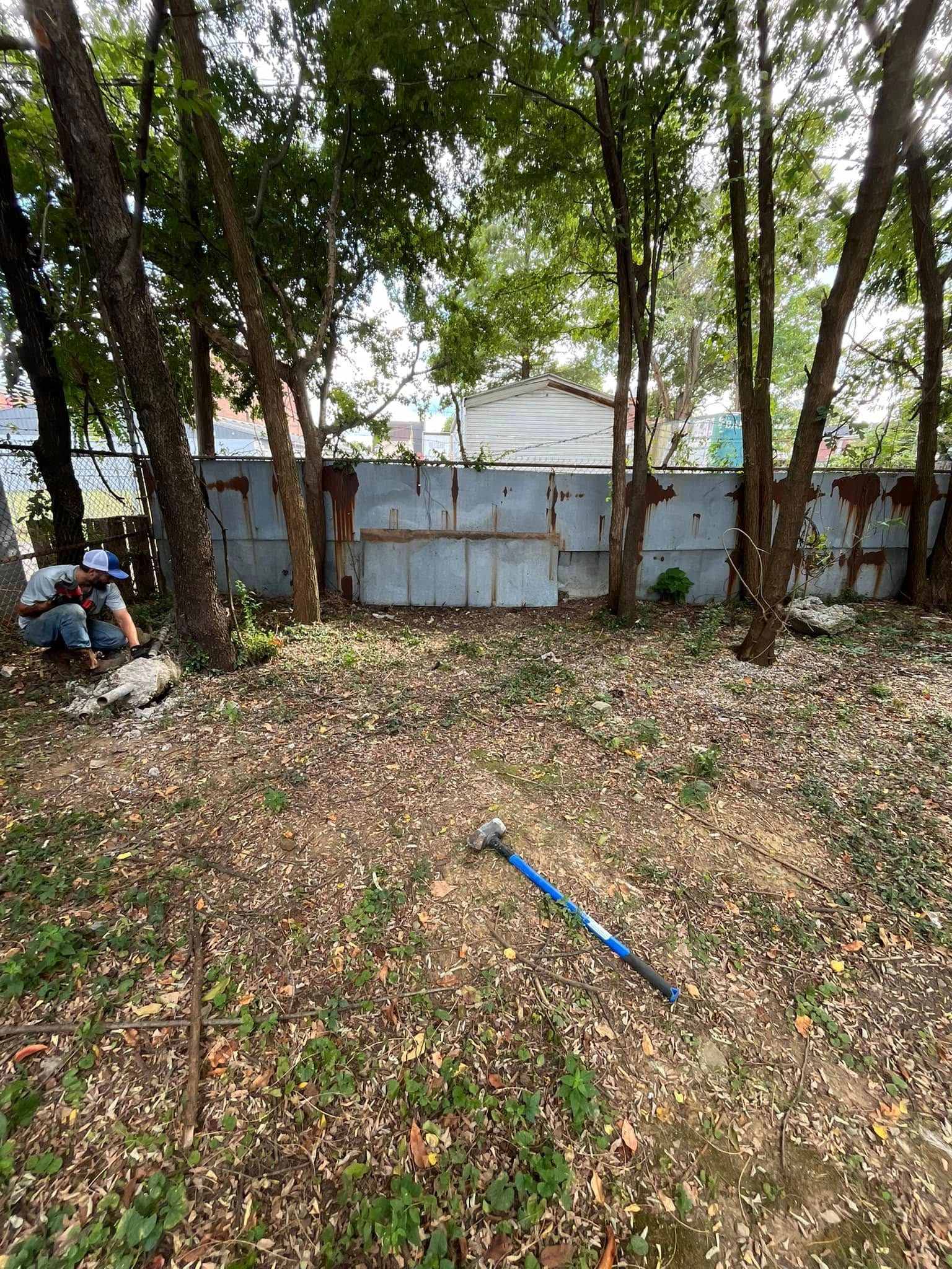 Yard with metal fence, trees, and fallen leaves. A person works on the left, blue handled tool lies in the foreground.