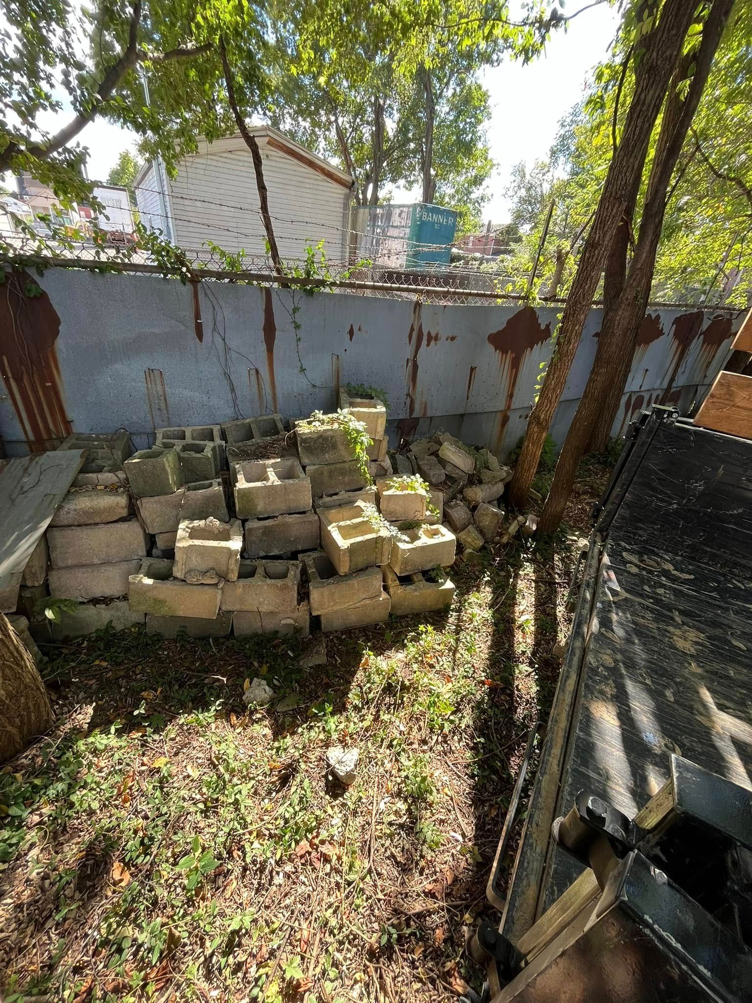 Pile of concrete blocks in a backyard, rust-stained metal fence and trees in background.