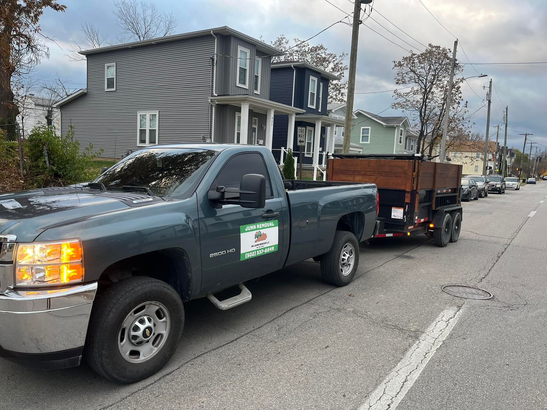 Dark gray pickup truck with trailer parked on a street in front of houses; cloudy day.