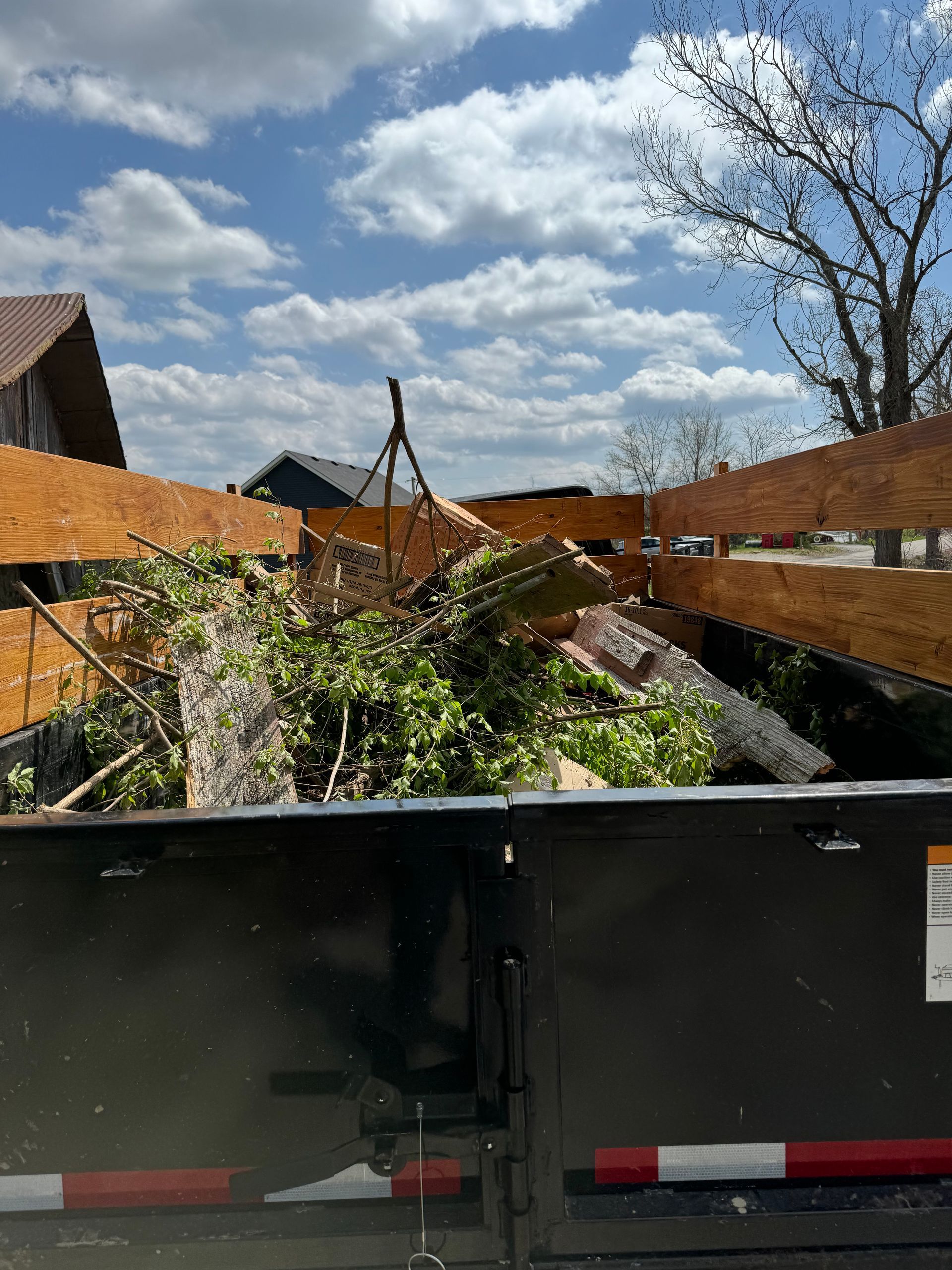 Black dumpster filled with tree branches and debris against a cloudy sky.