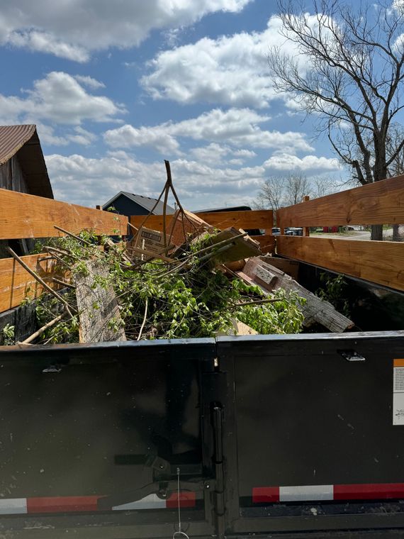 Black dumpster filled with tree branches and debris against a cloudy sky.