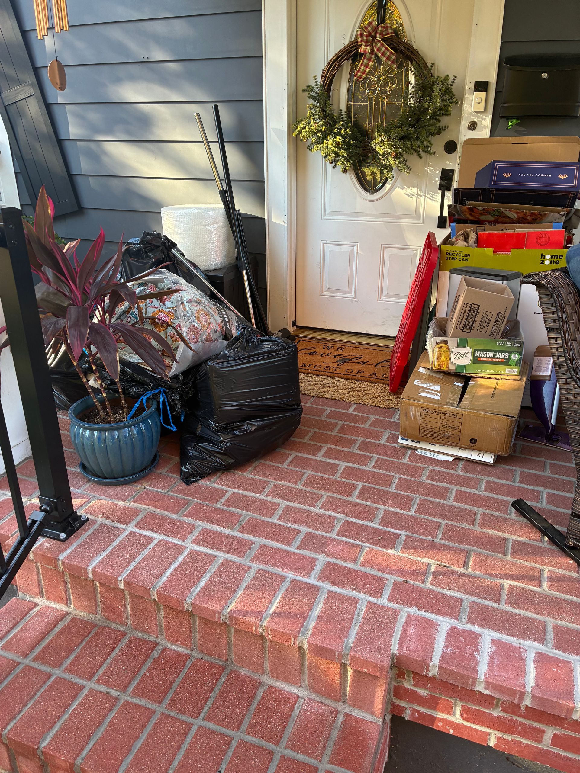 Red brick porch with clutter near a white door, including a trash bag, plant, and boxes.