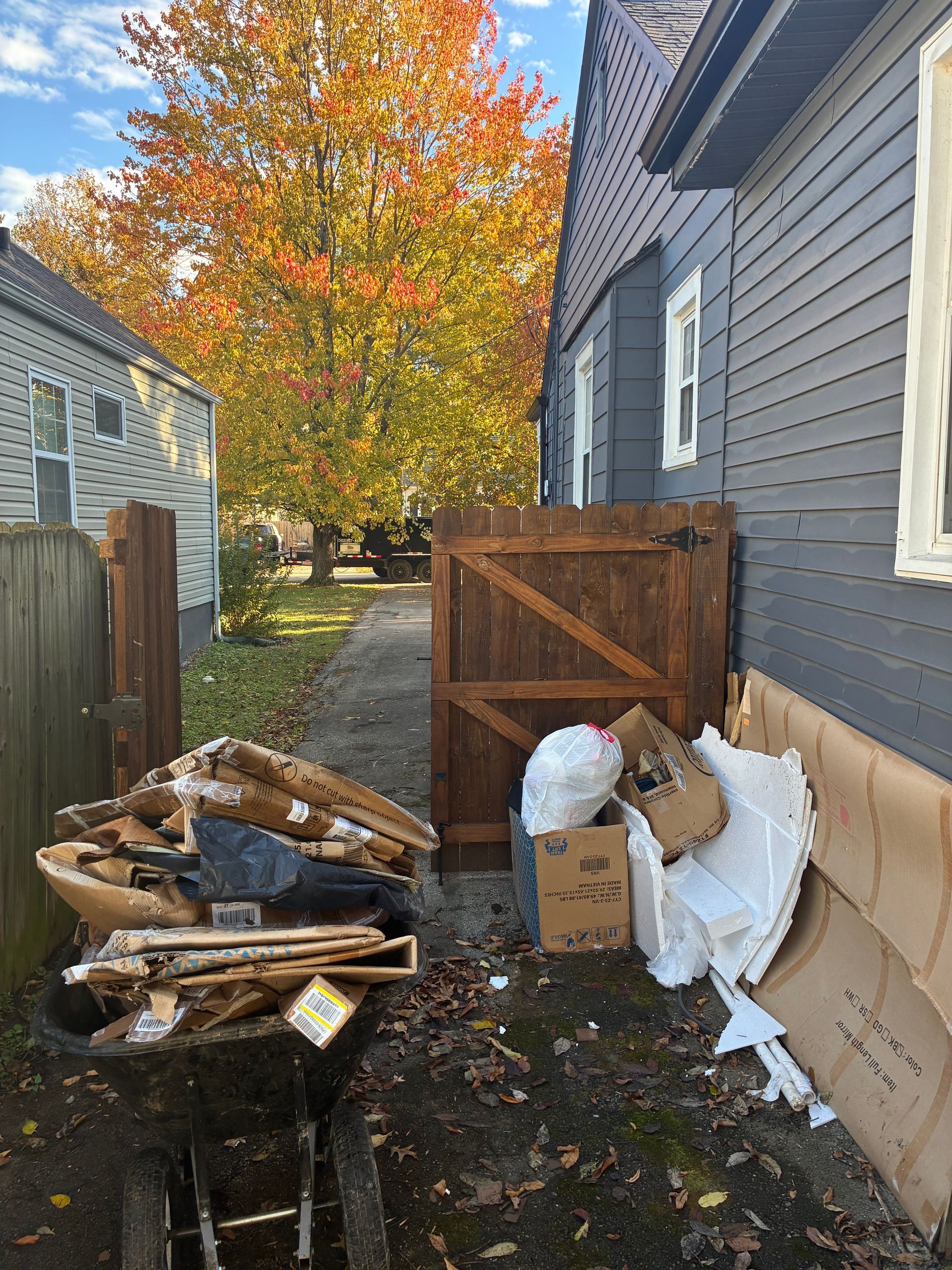 Pile of discarded cardboard, wood, and trash next to a wooden fence and gray house. Fall foliage in the background.