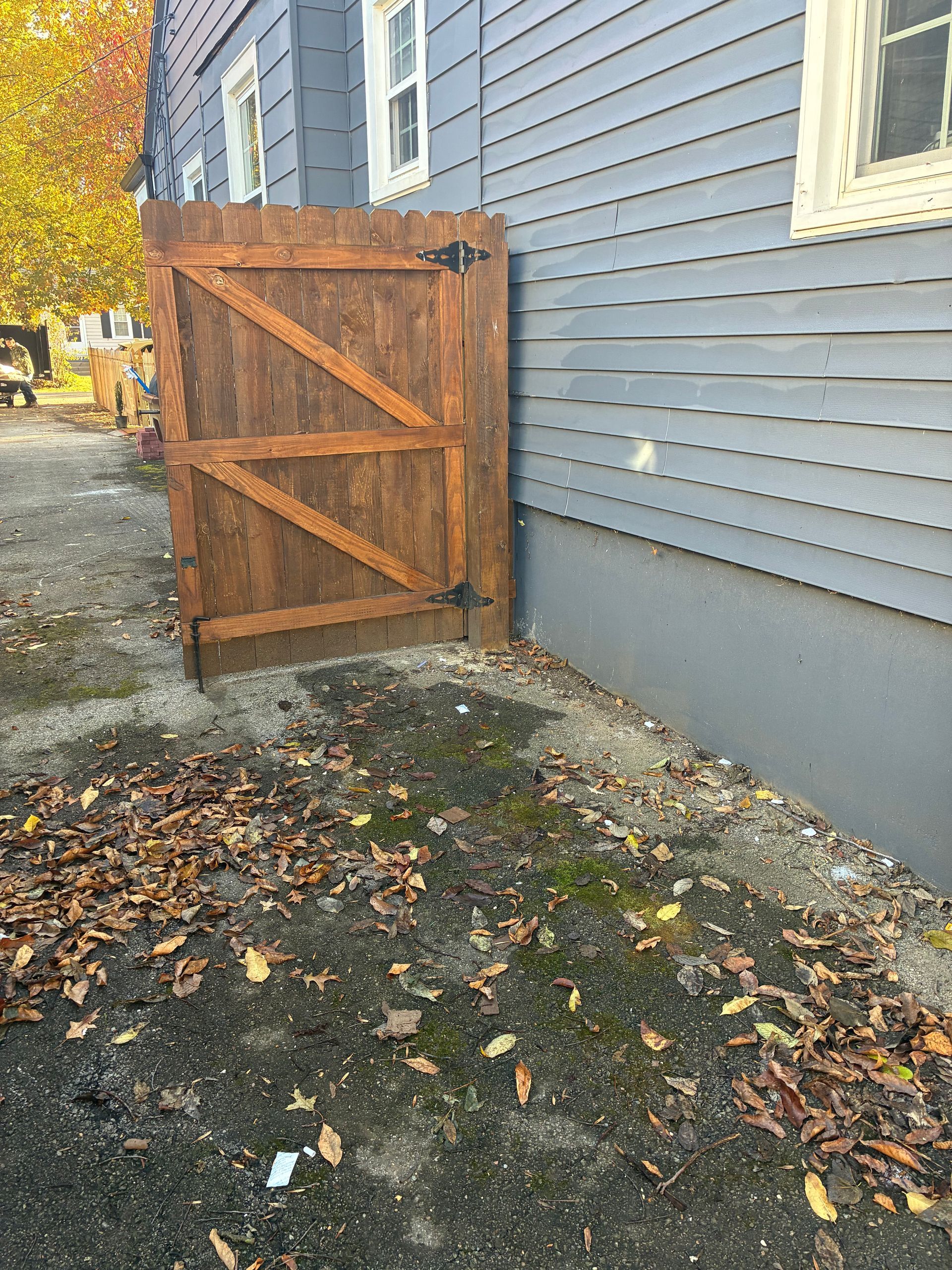Wooden gate, open, against a blue house with gray foundation. Brown leaves on the ground.