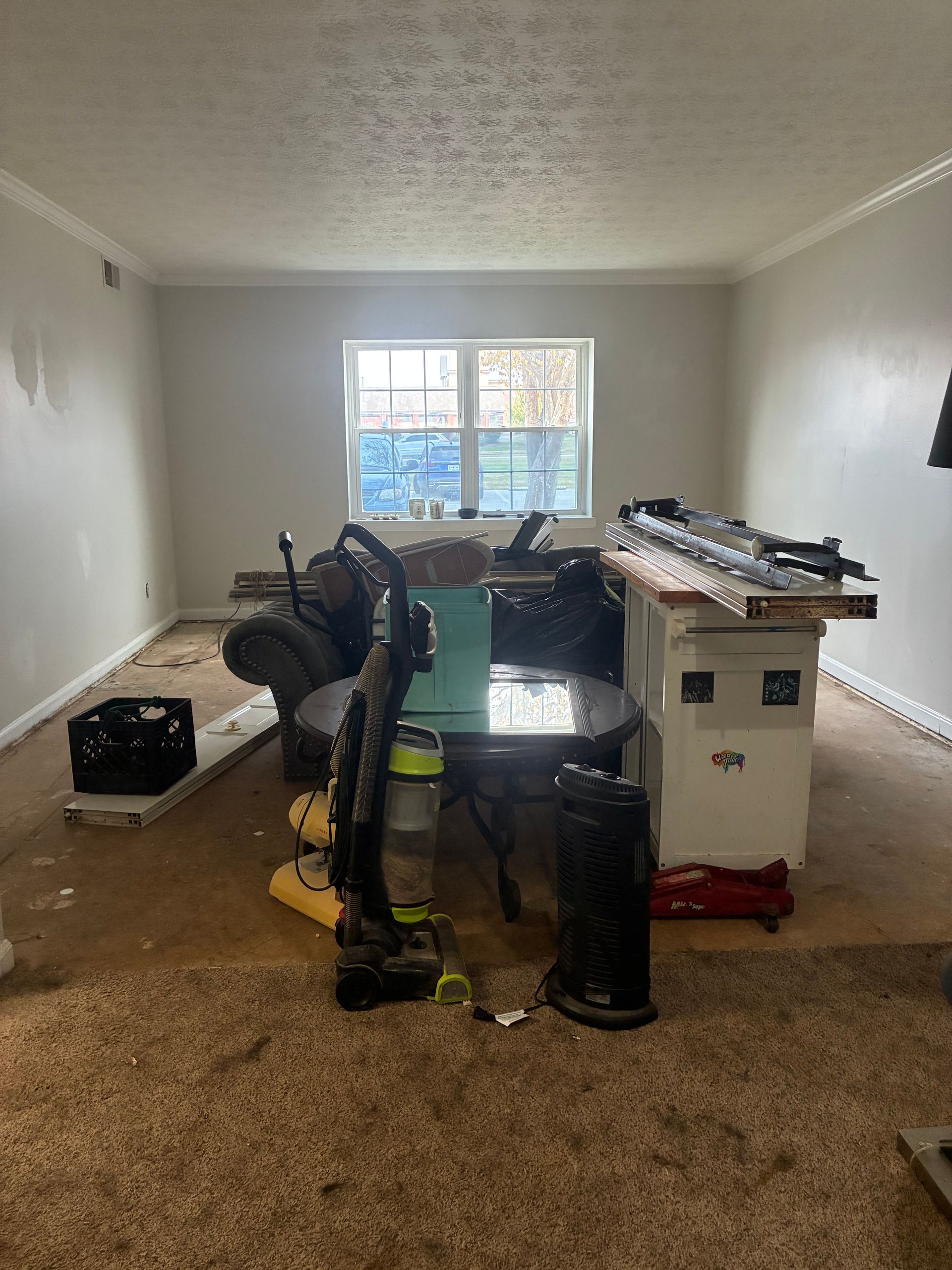 An empty room with tools and equipment piled on a table and floor, centered in front of a window.