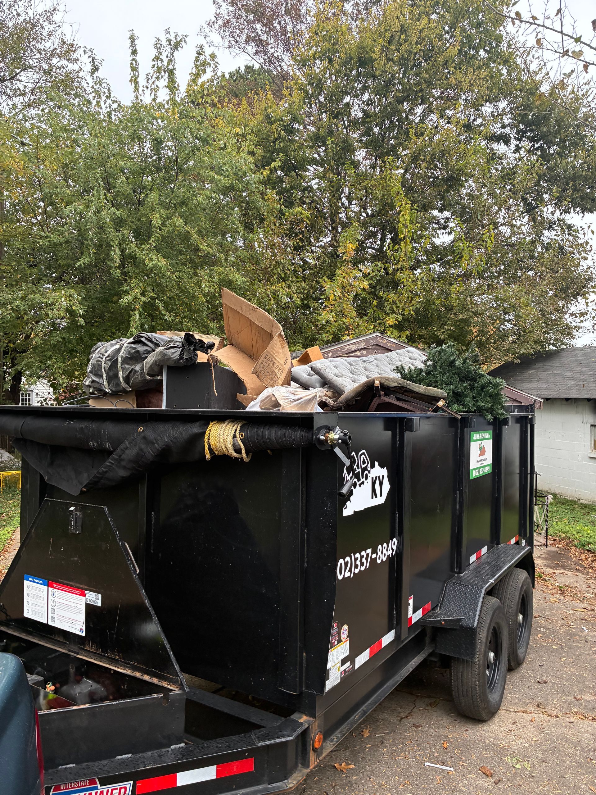 Black dumpster trailer filled with debris, parked outside a house, with trees in the background.
