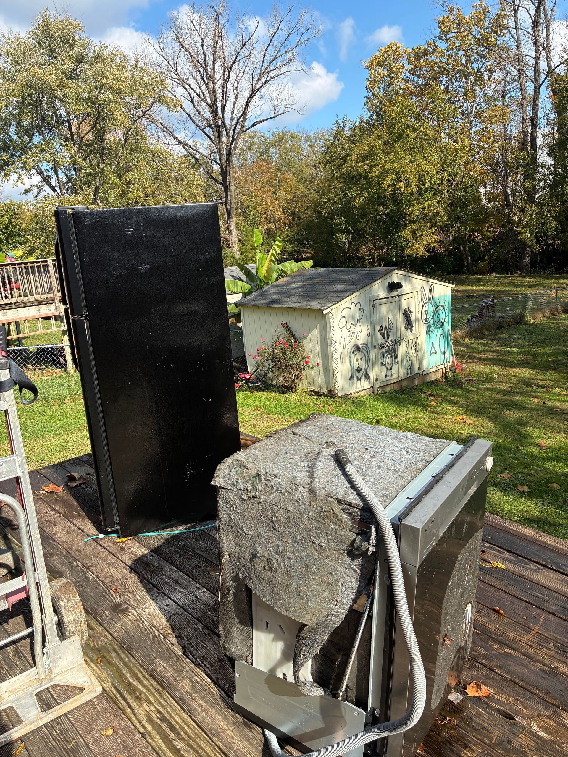 Refrigerator parts on a wooden deck in front of a shed, under a blue sky.