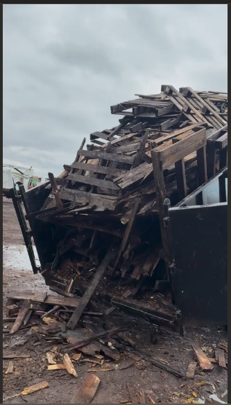 A large container overflowing with weathered wooden debris, likely construction waste, set outdoors under a cloudy sky.
