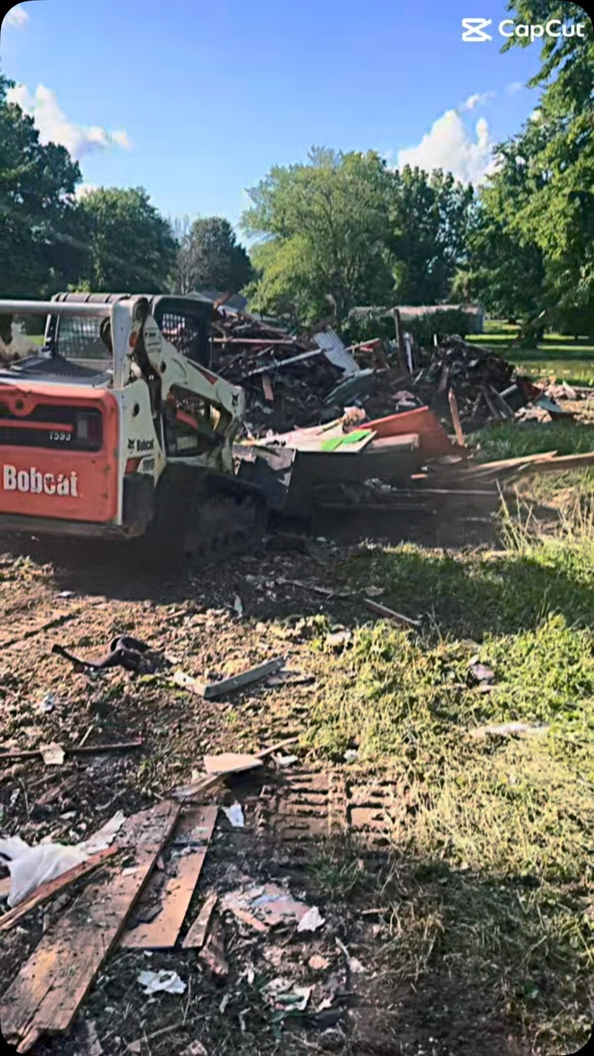 Bobcat skid-steer loader moving debris in a grassy area with trees under a blue sky.