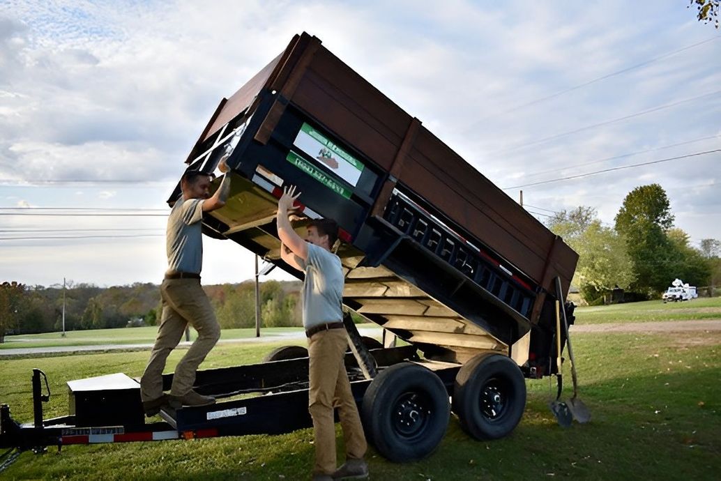 Two people lift the bed of a black and brown dump trailer on a grassy field.