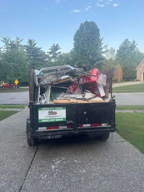 A black trailer overflowing with construction debris sits on a driveway in front of a house, with green grass and trees in the background.