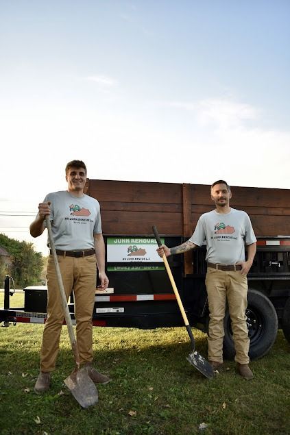 Two men in matching shirts stand with shovels in front of a trailer.
