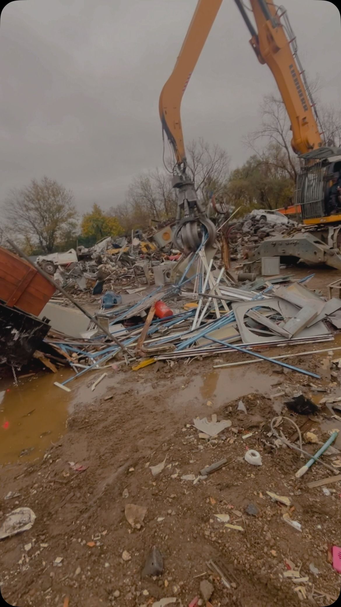 An excavator claw crushing metal debris in a muddy scrap yard under an overcast sky.