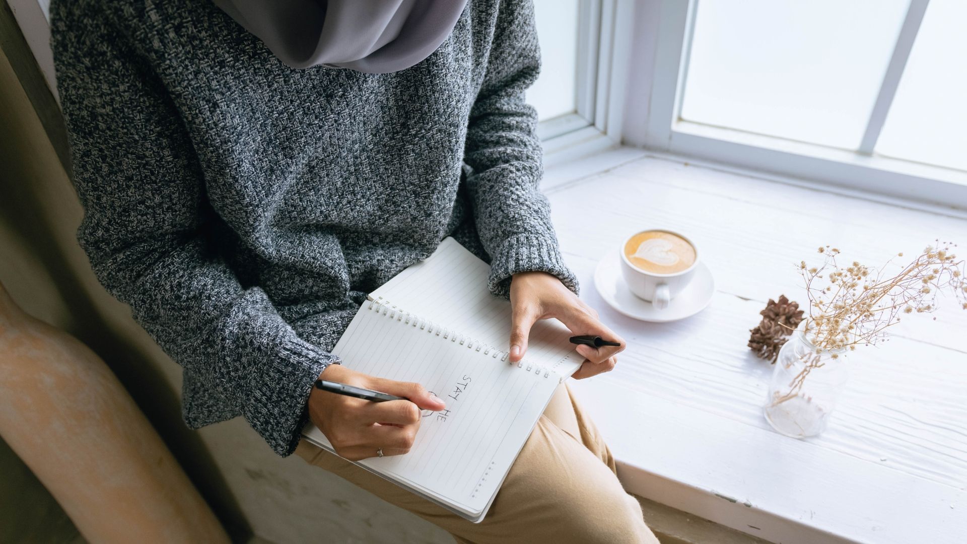 Woman in grey sweater and hijab writing in a notebook on a windowsill with a coffee and dried flowers.