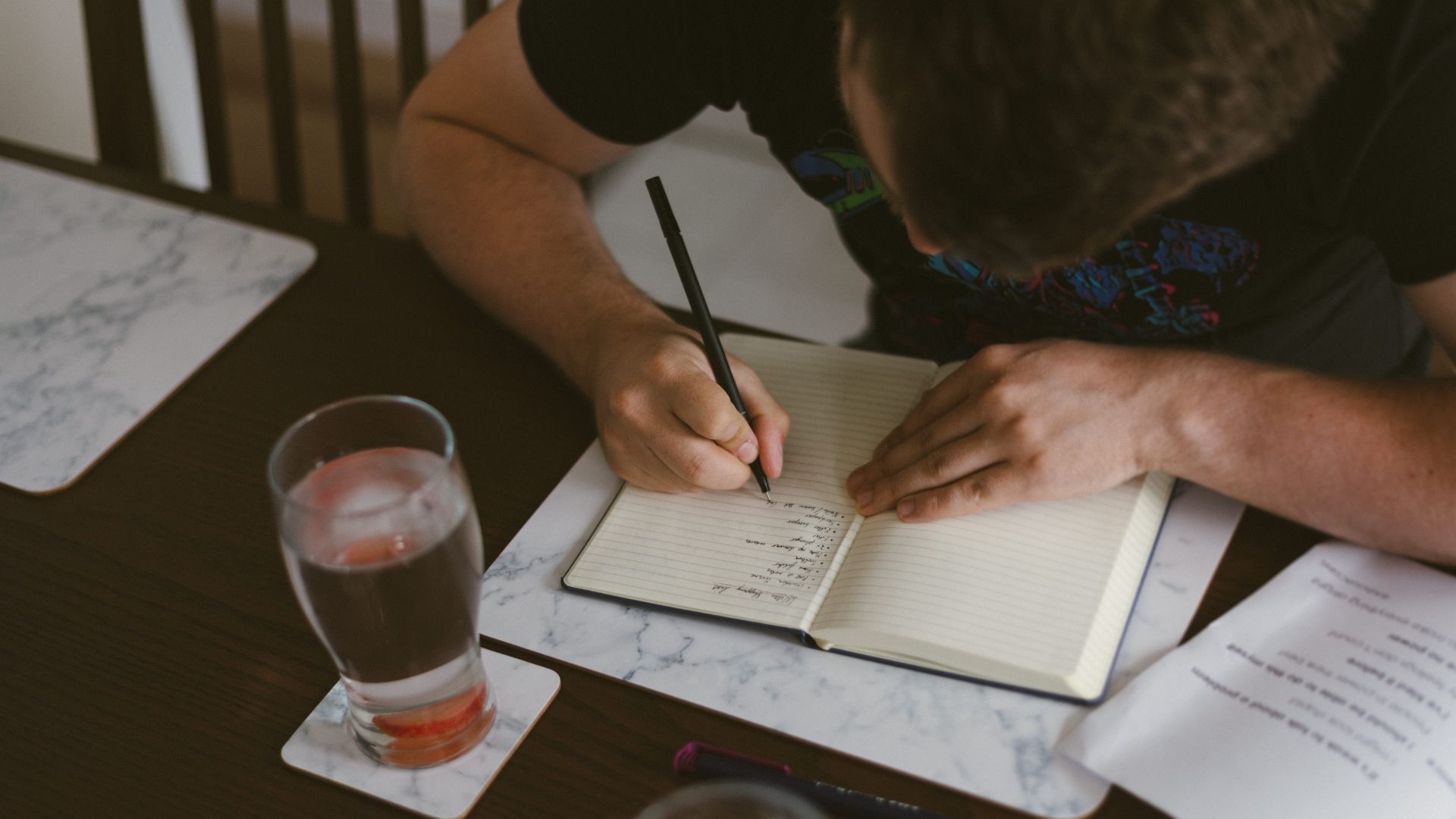 Man writing in notebook at a table with drink and papers.