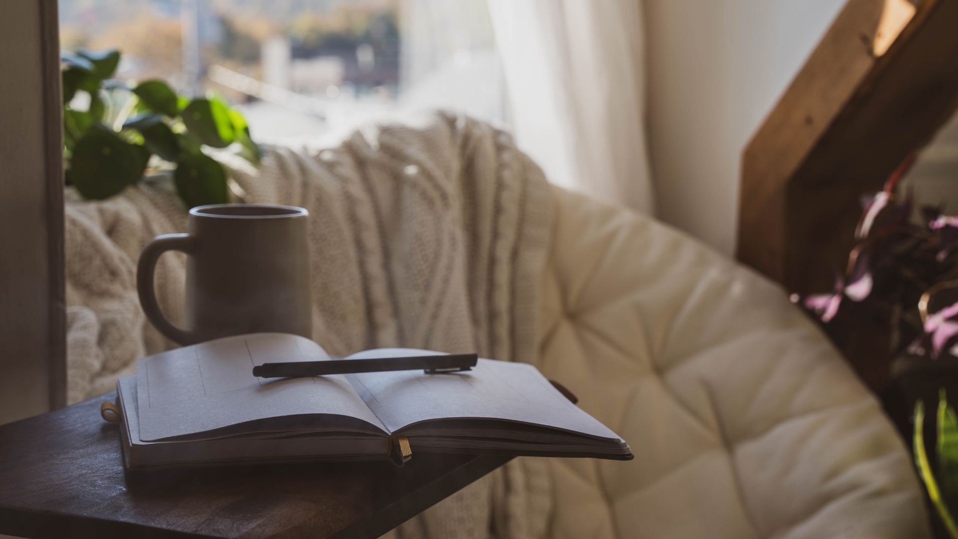 Cozy reading nook: open notebook and pen, mug of coffee, soft chair by window with plants.