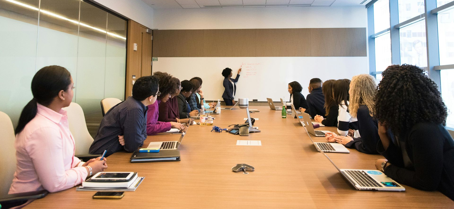 People in a meeting room, a person presenting at a whiteboard. They are sitting around a conference table with laptops.