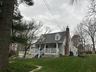 A gray house with a porch, chimney, and dormers sits on a green hill under a cloudy sky.