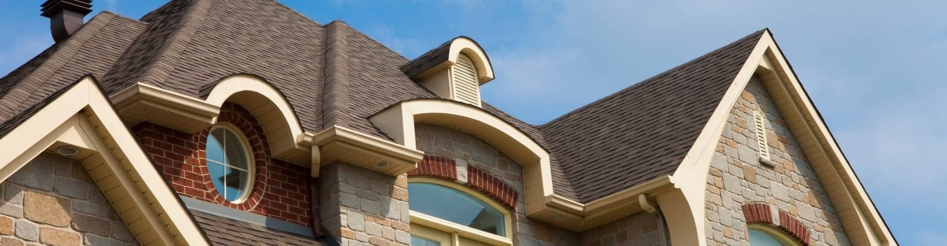 A stone house with a brown roof and dormers against a blue sky.
