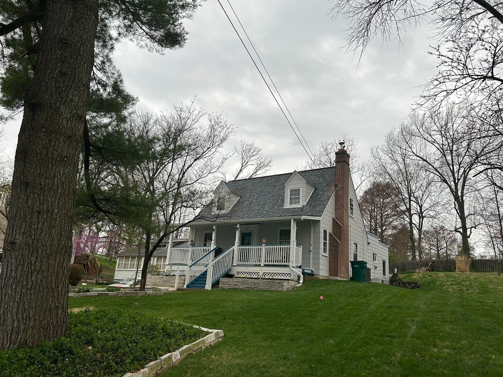 White house with porch, chimney, and dormers under a cloudy sky. Green grass, trees.