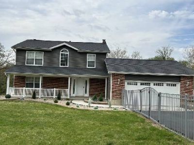 Two-story house with gray siding, brick, a porch, and a garage; green lawn and cloudy sky.