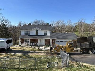 House with tree removal in progress; worker operating a wood chipper.