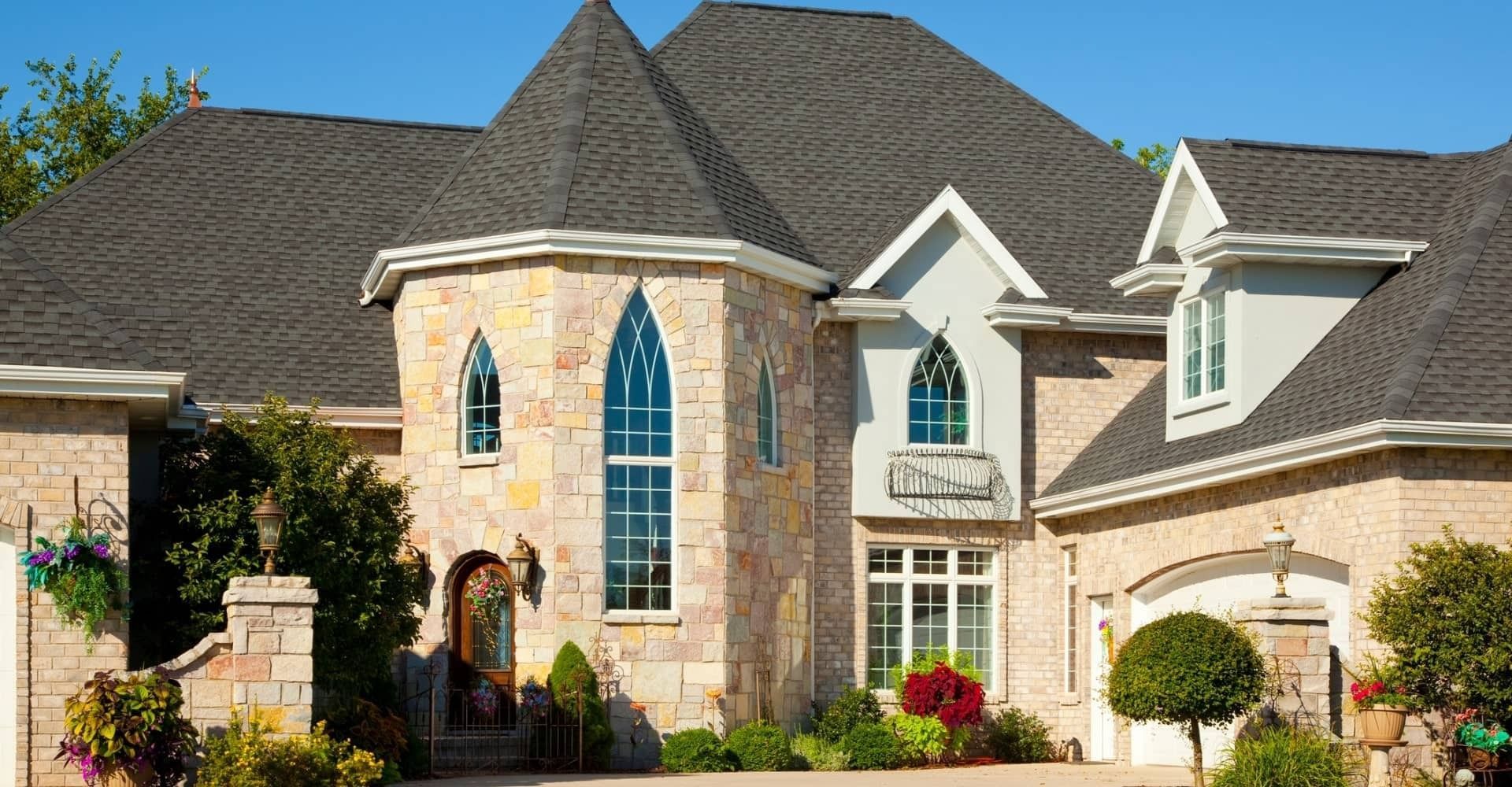 Stone and brick house with dark gray roof, ornate windows, and lush landscaping.