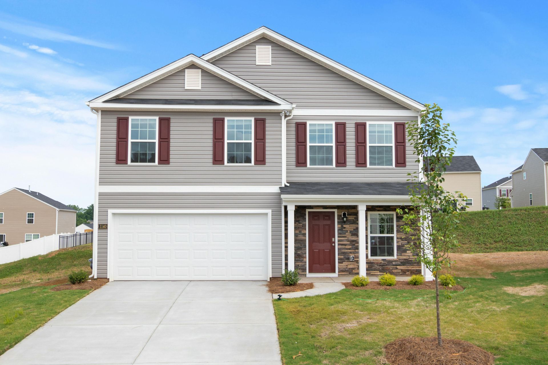 Two-story gray house with red shutters and a white garage door, on a green lawn under a blue sky.