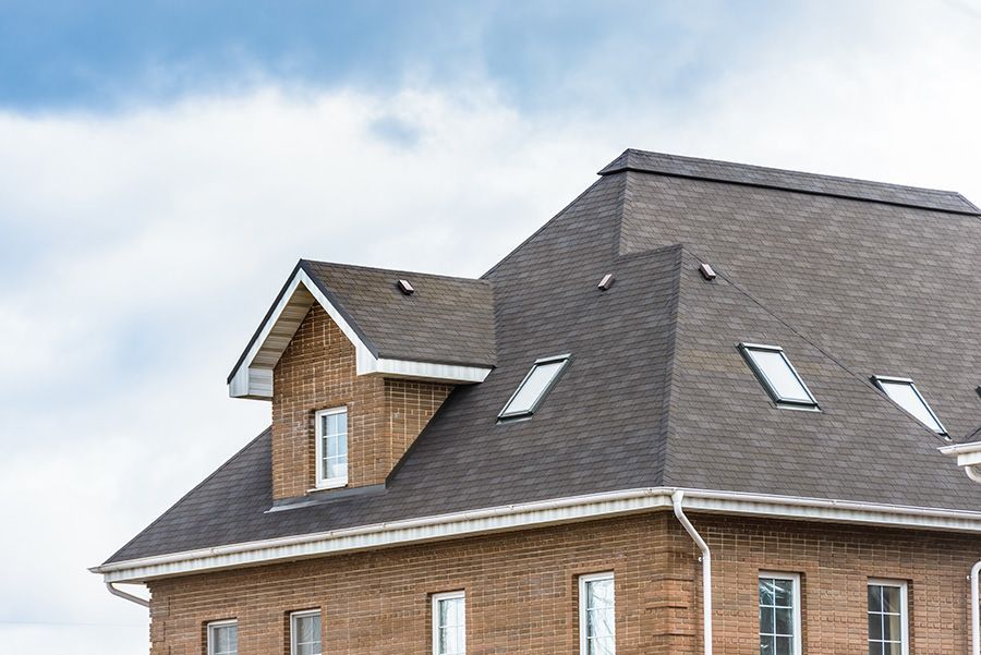 Brick house with brown shingle roof, dormer, and white trim against a cloudy sky.