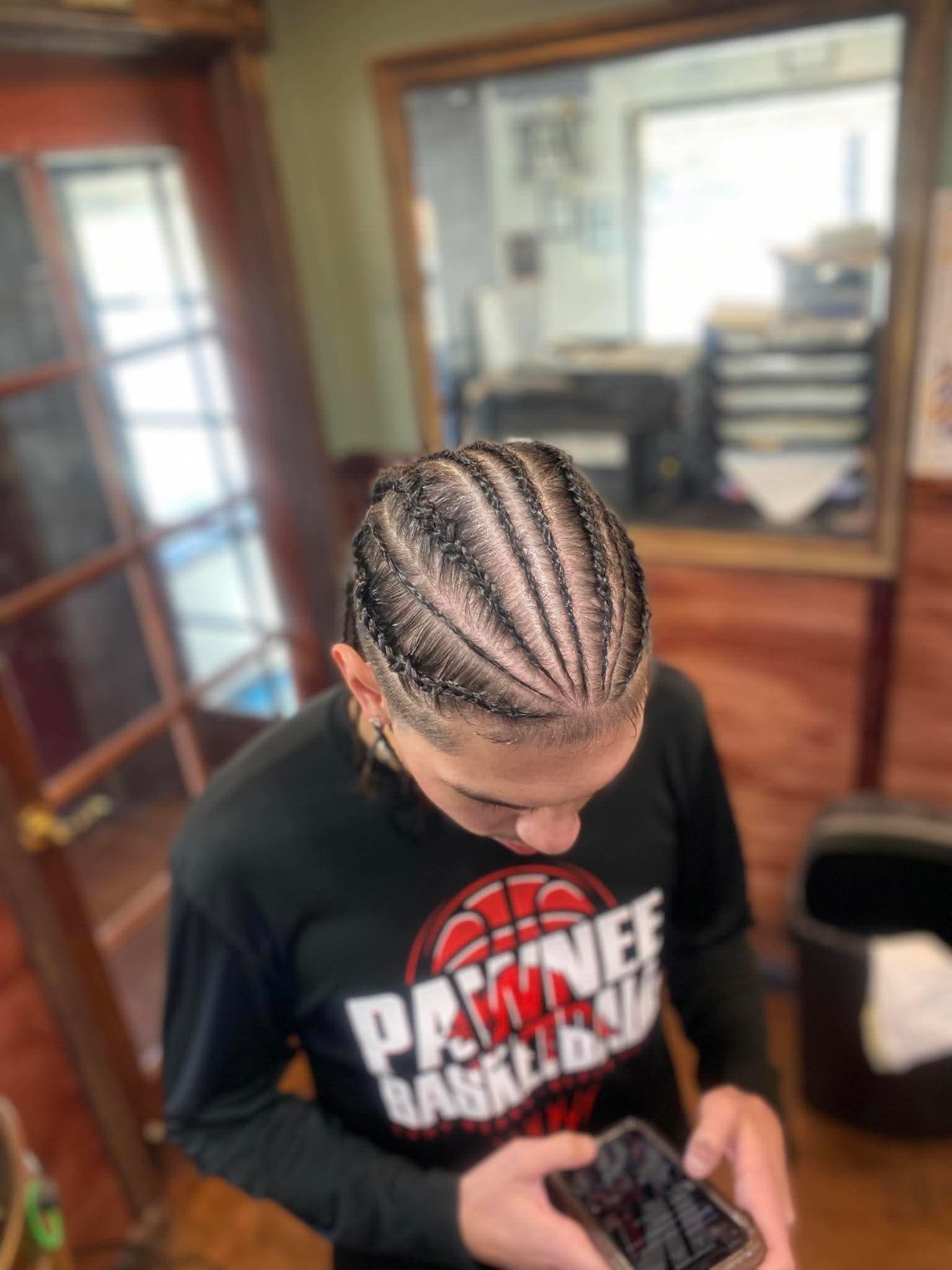 A person with cornrows styled in straight back braids, wearing a long-sleeved black Pawnee Basketball shirt indoors.