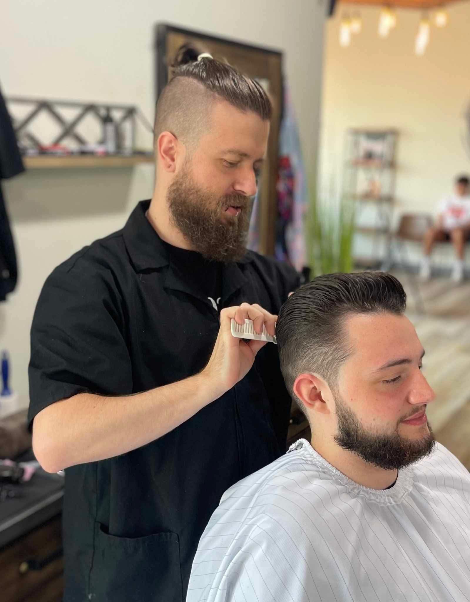 A barber with a beard and top-knot haircut uses a comb to style a client's hair in a brightly lit shop.