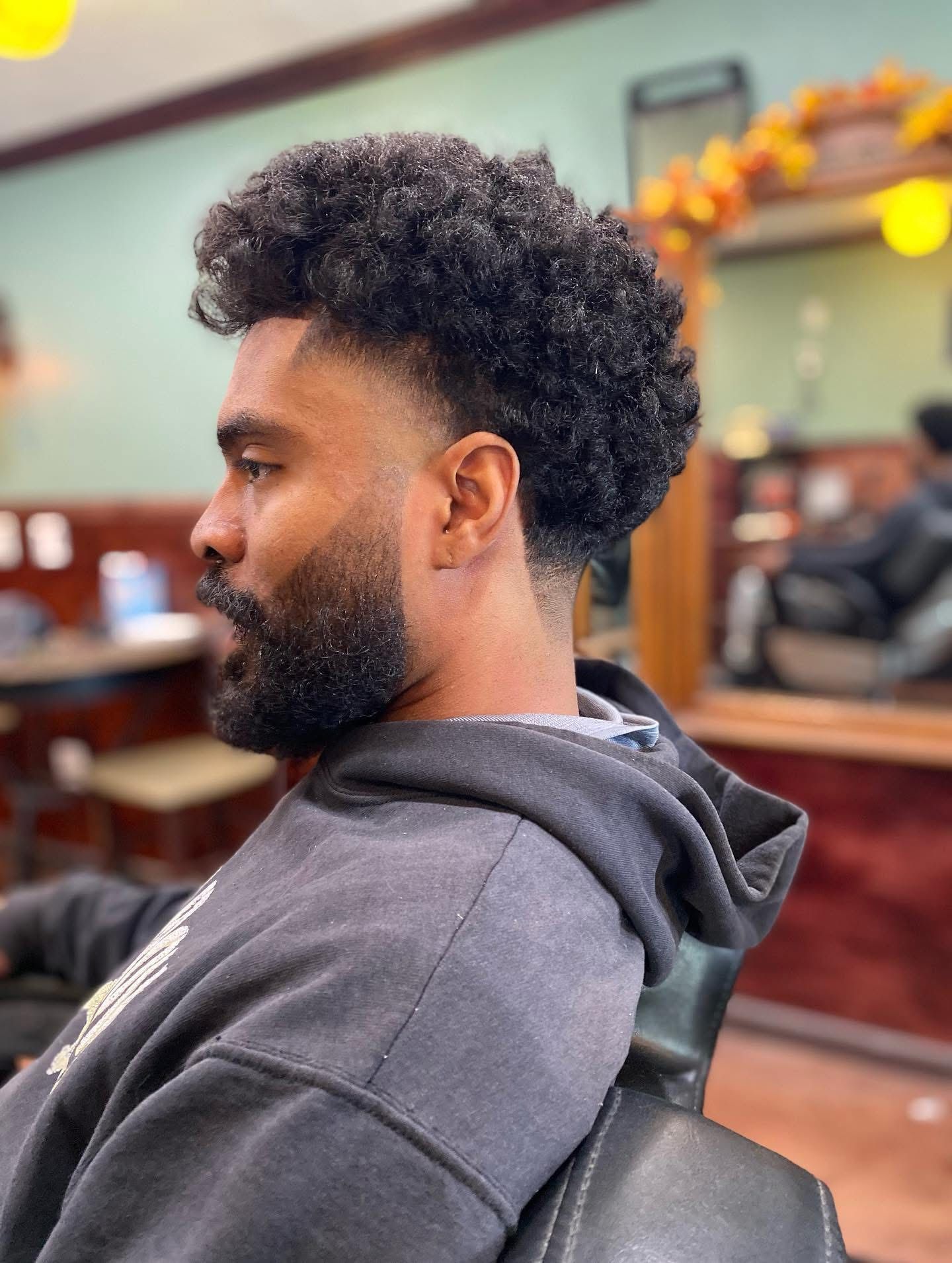Side profile of a person with a textured afro haircut and a faded beard, sitting in a barbershop chair.