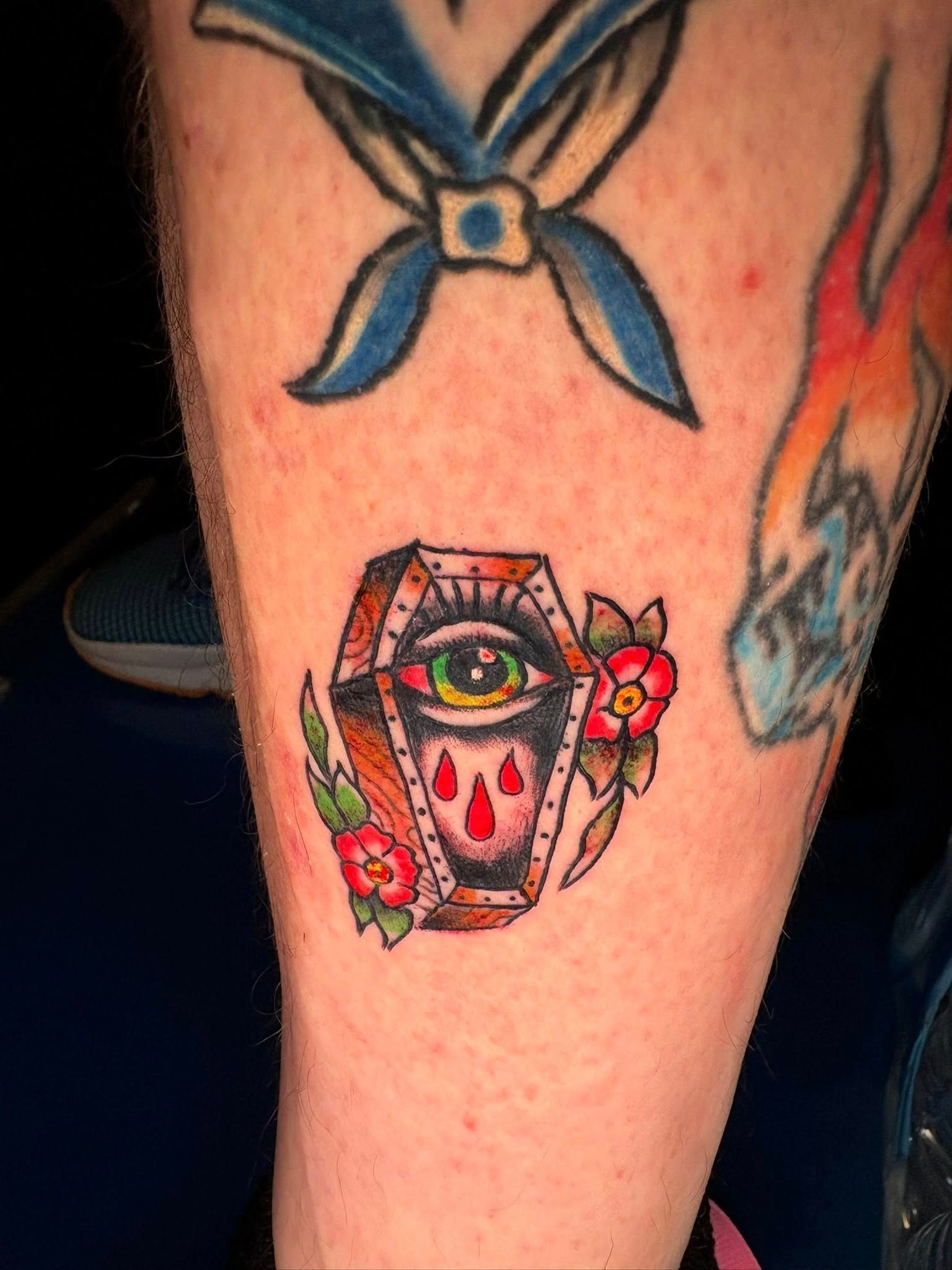 A colorful traditional-style tattoo of a coffin containing a weeping eye, decorated with red flowers, on a person’s leg.