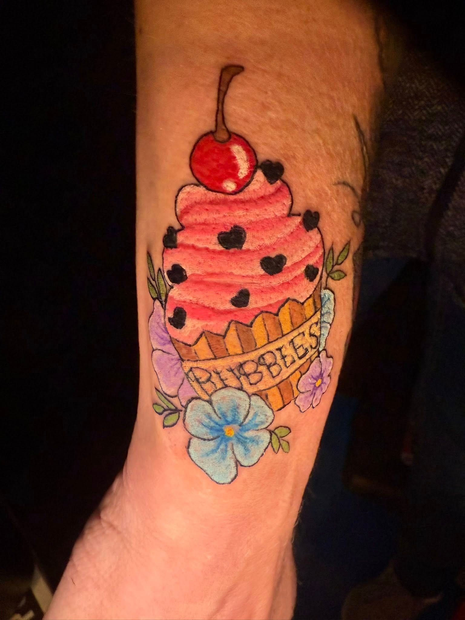 A colorful tattoo on an arm of a cupcake topped with a cherry and heart-shaped sprinkles
