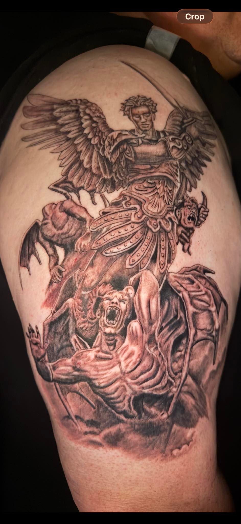 A black-and-grey tattoo on an upper arm depicting an angel standing over a fallen, horned figure with large wings.