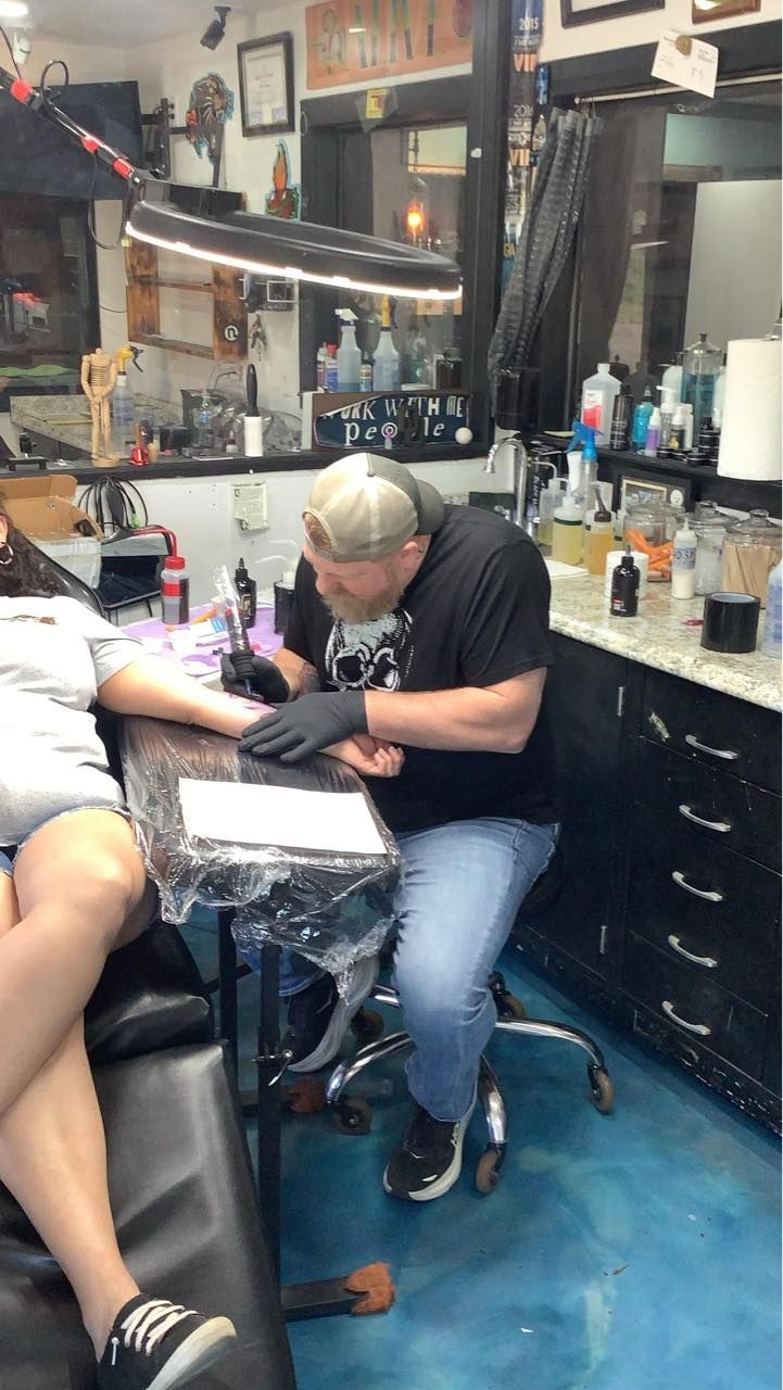A tattoo artist in a black shirt and cap works on a client’s forearm in a brightly lit studio with black cabinets.