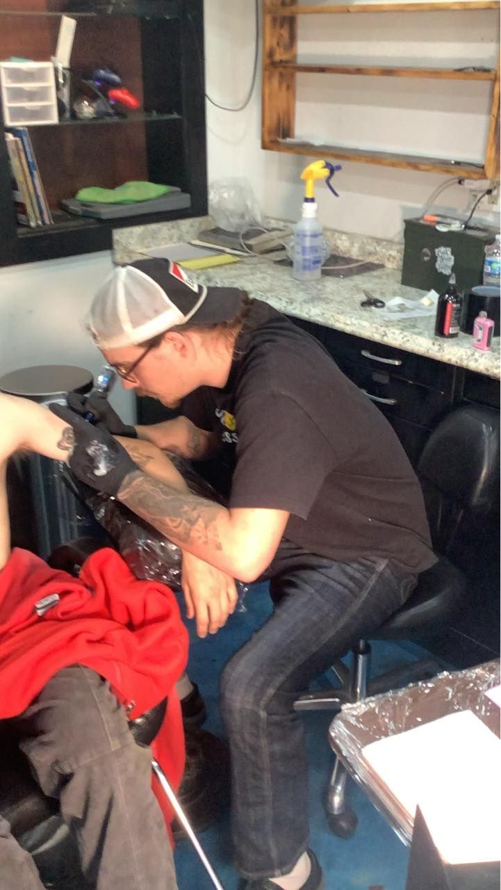 A tattoo artist in a cap and black shirt focuses on tattooing a client's arm in a well-equipped studio.